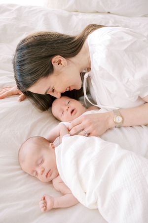 A tender moment captured as a mother lies with her twin babies on white bedding.