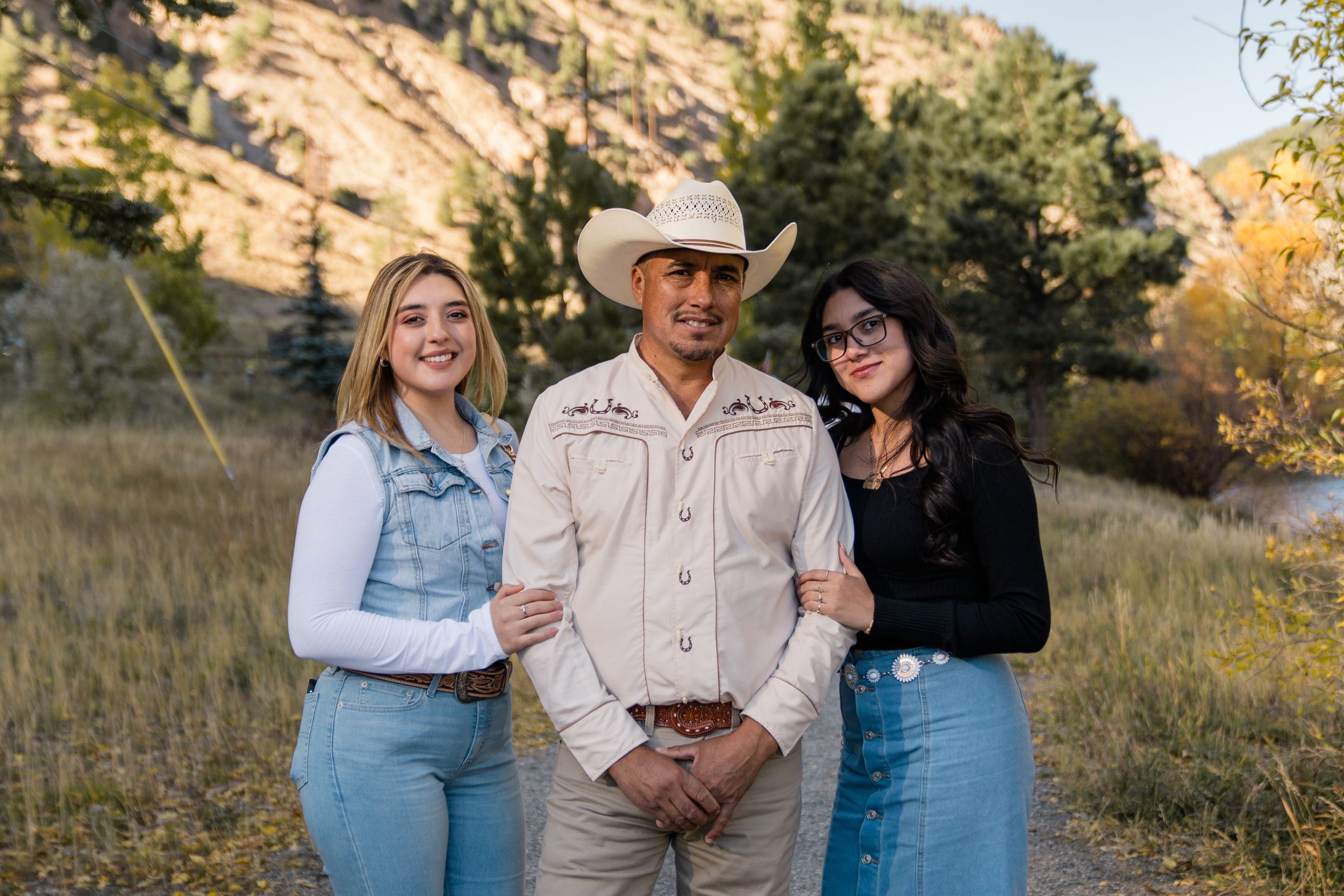 Three people in casual attire stand together in a golden field during sunset creating a warm family portrait.