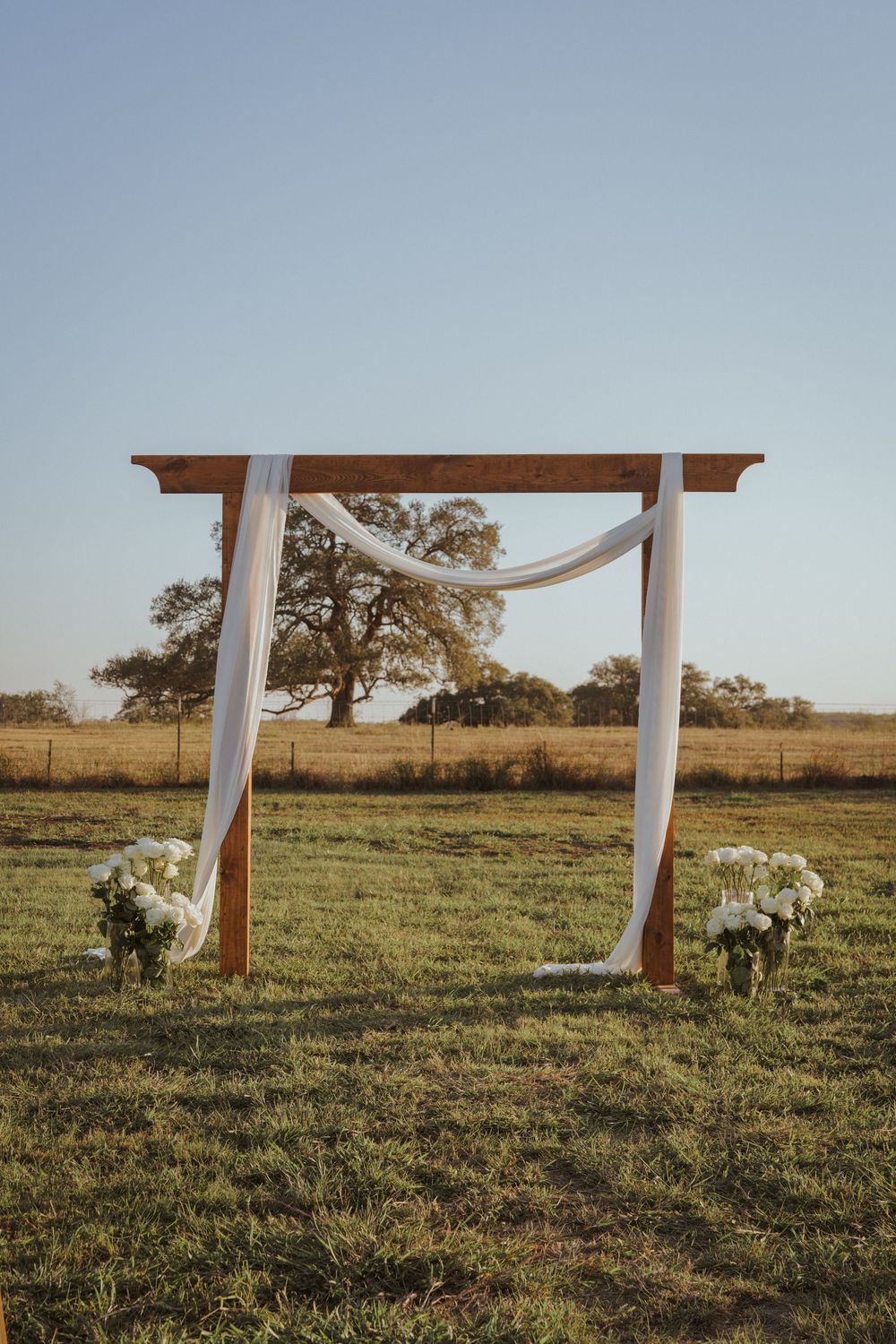Wedding arch with white fabric and floral arrangements in a rustic outdoor setting.