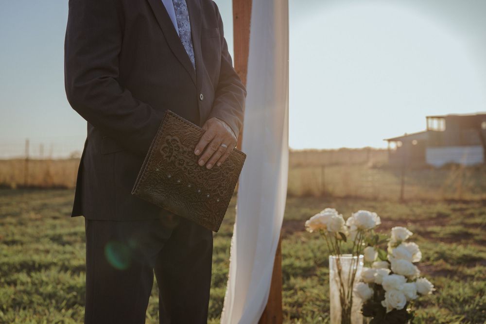 Rustic outdoor wedding ceremony setup in a field at sunset with wooden arch and white floral arrangements.