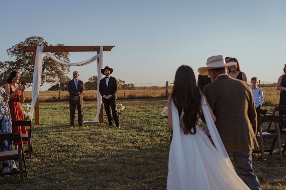 Wedding guests gathered for outdoor ceremony under wooden arch in rustic countryside setting.