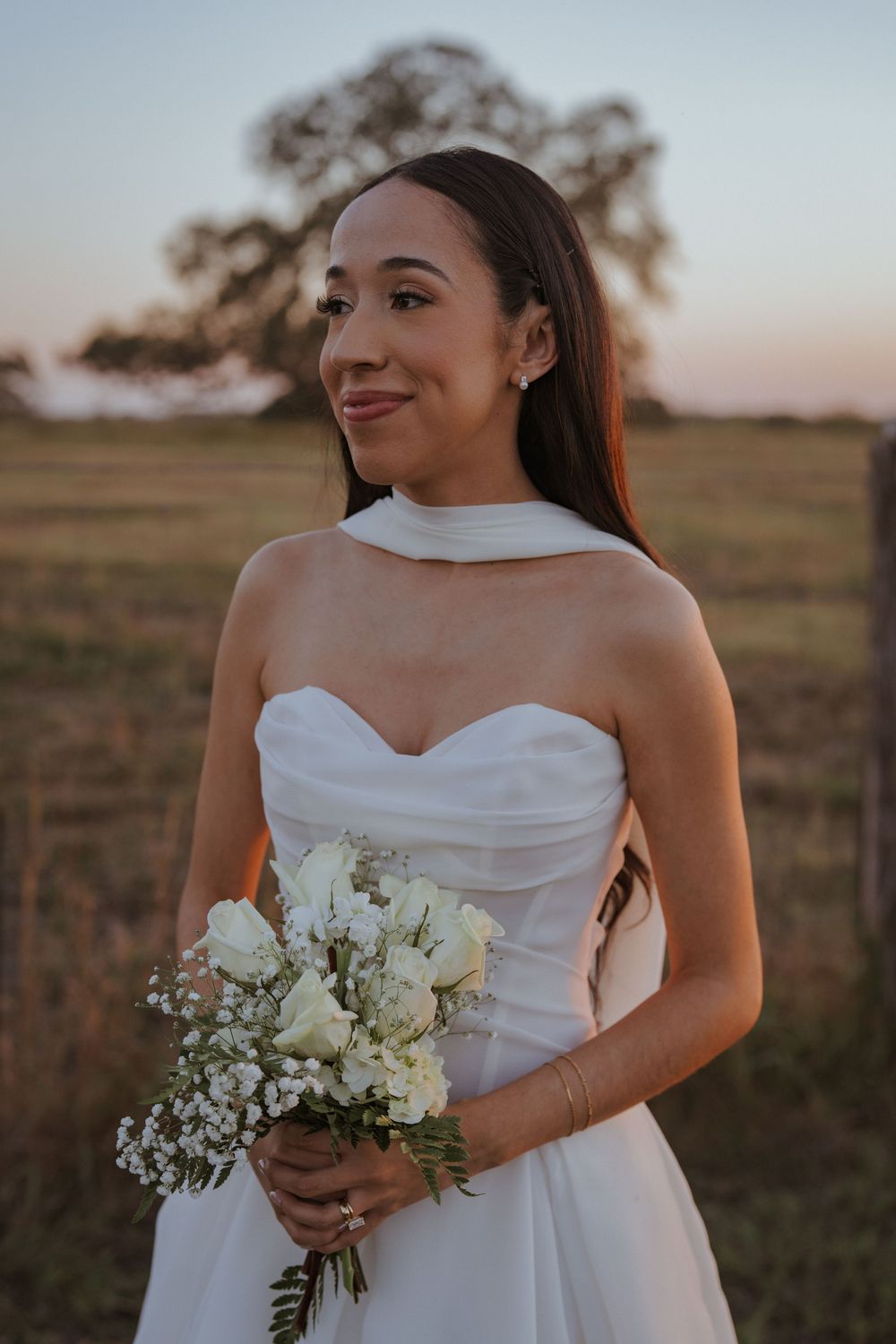 Bride in white dress holding delicate bouquet of white flowers during sunset outdoor photoshoot.
