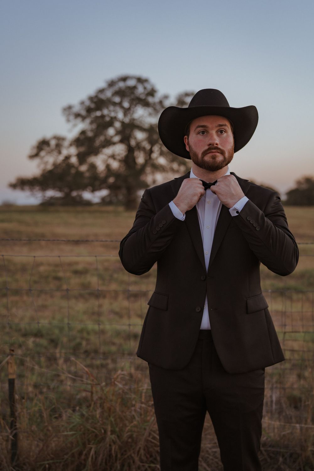 Person in black suit and cowboy hat adjusting their tie in a field at dusk.