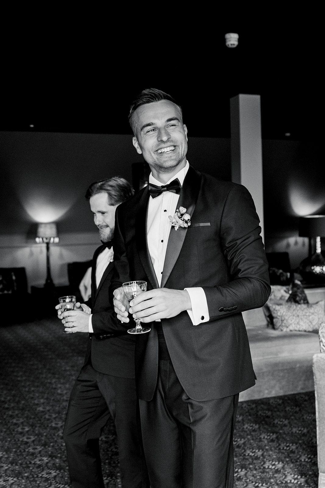 Wedding party members in black tuxedos share drinks and laughter during an evening celebration.