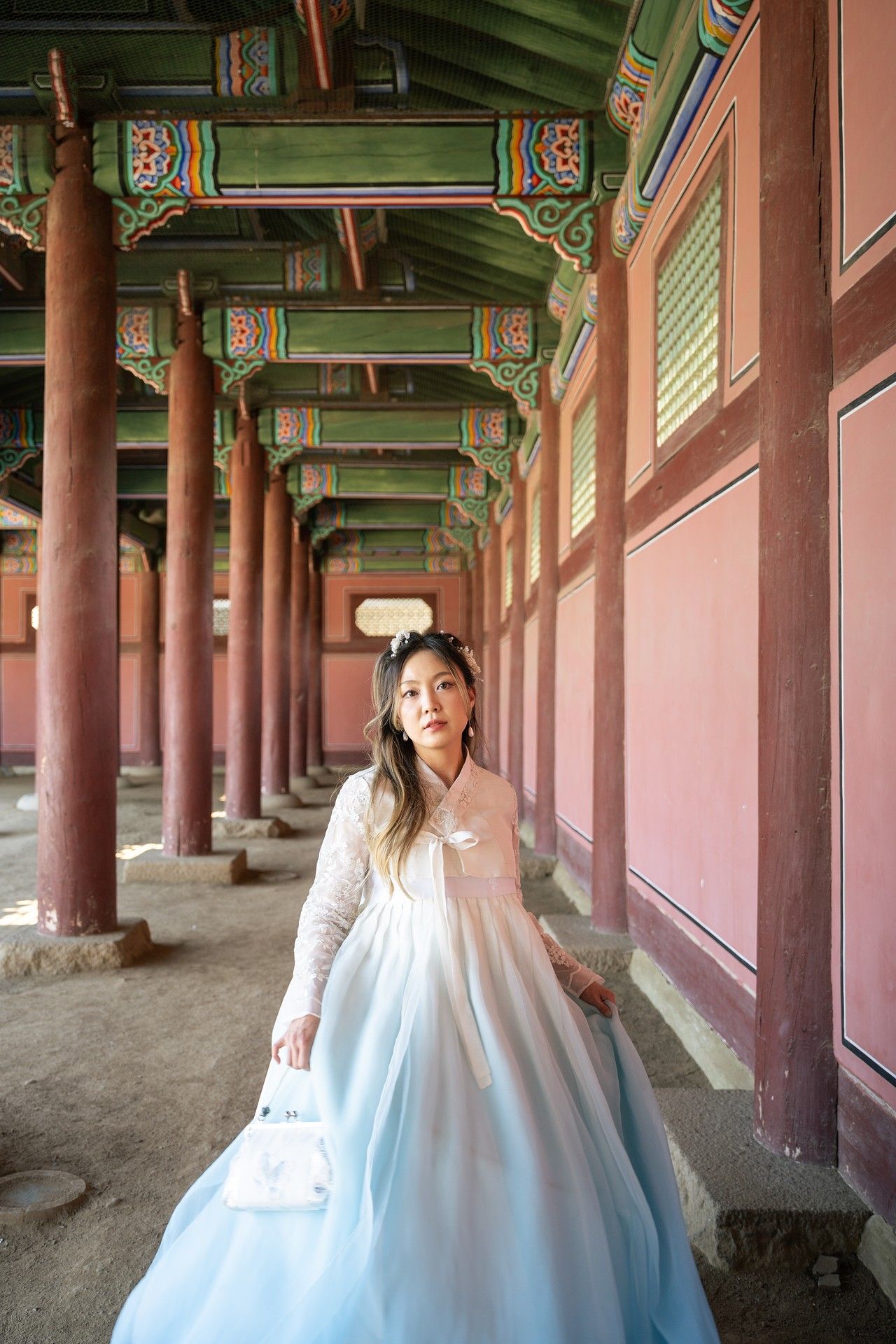 Traditional Korean hanok corridor with painted wooden beams and columns features someone in a flowing white hanbok dress.