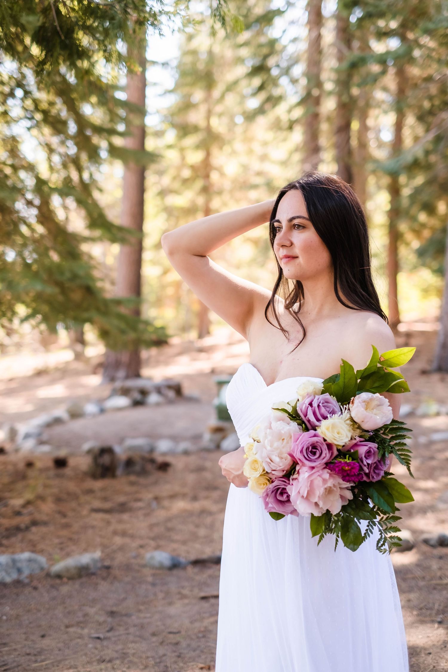 A bride in a white dress poses gracefully with a colorful bouquet of flowers in the tree line on the banks of lake tahoe ca