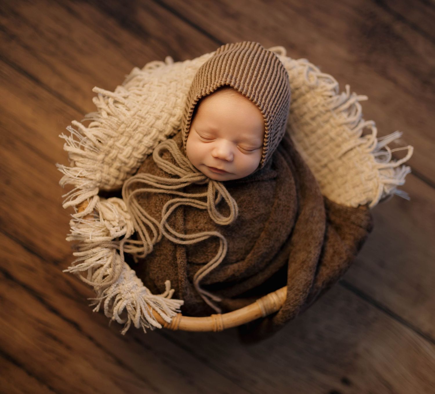A newborn baby sleeps peacefully in a cozy brown knit wrap on a wooden surface with fringe blanket details.