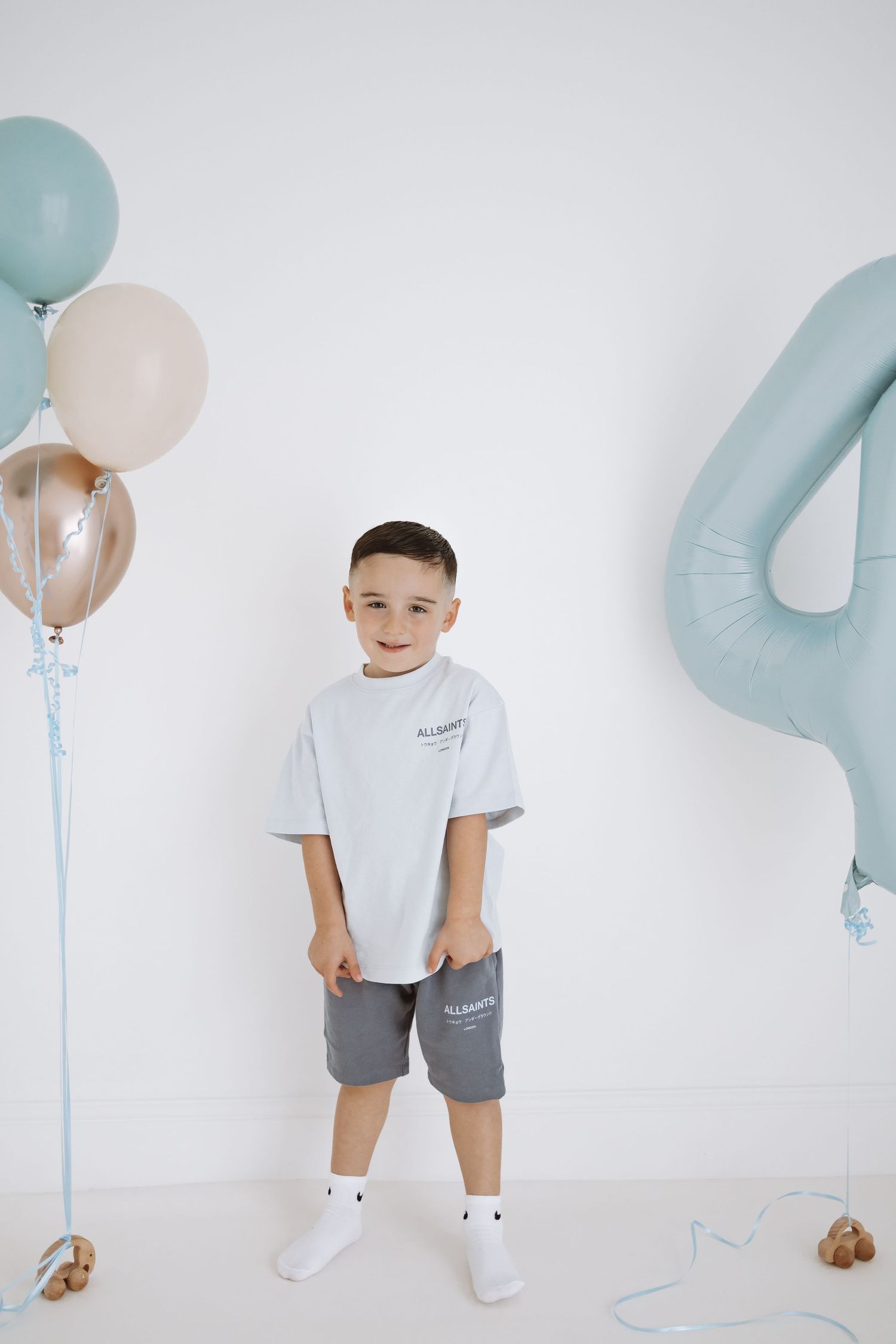 First birthday celebration setup with blue and gold balloons, white backdrop, and baby wearing ONE shirt sitting and smiling.