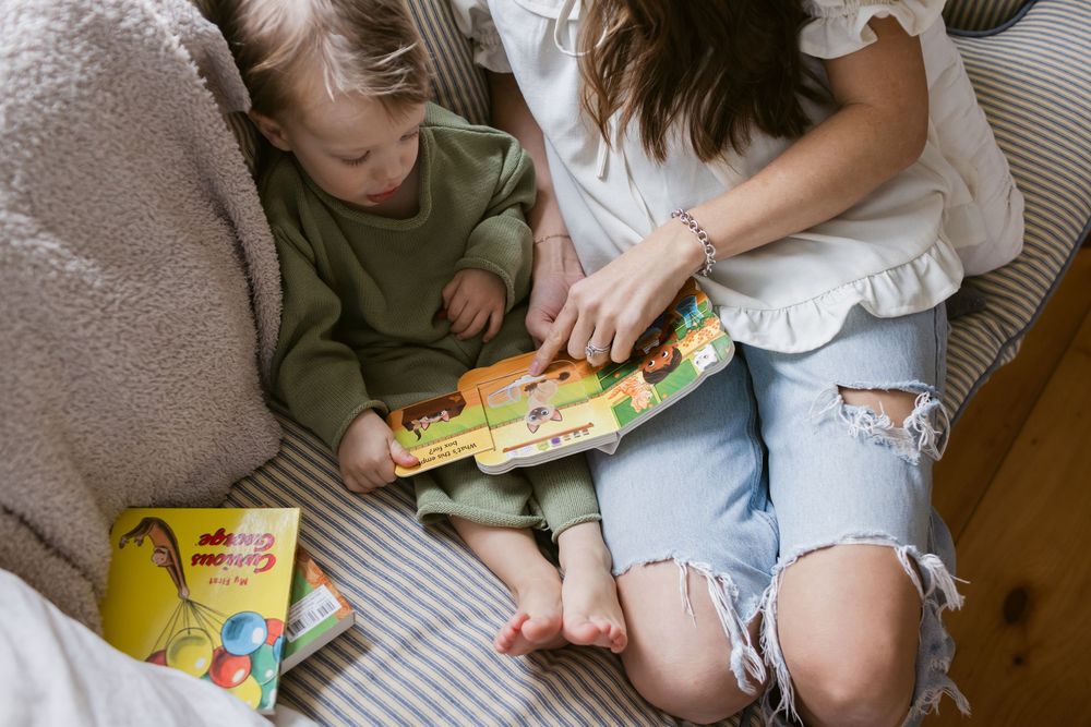 An above angle of a mother with ripped jeans and white peasant shirt sits next to her toddler son wearing a green sweater outfit reading
