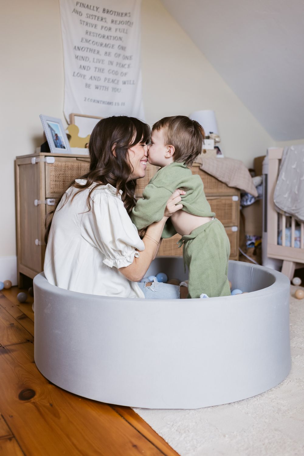 A toddler son kisses his mother on her nose while playing in foam ball pit in his light and airy bedroom for family photos in Harvard, MA