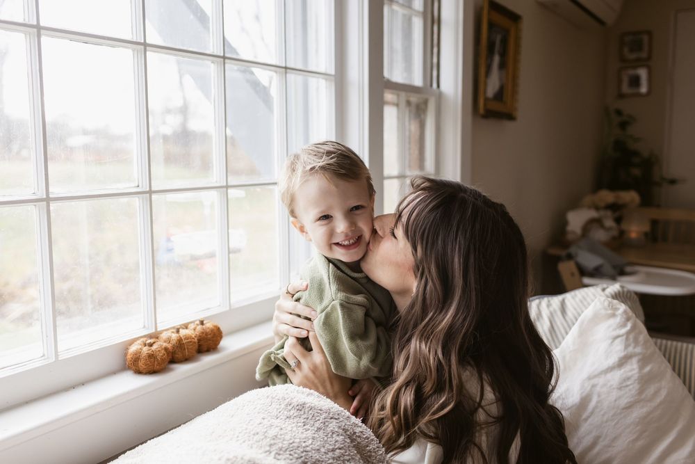 A mother kisses her toddler son while sitting together by large windows in a bright living room during a mommy and me shoot in Harvard, MA