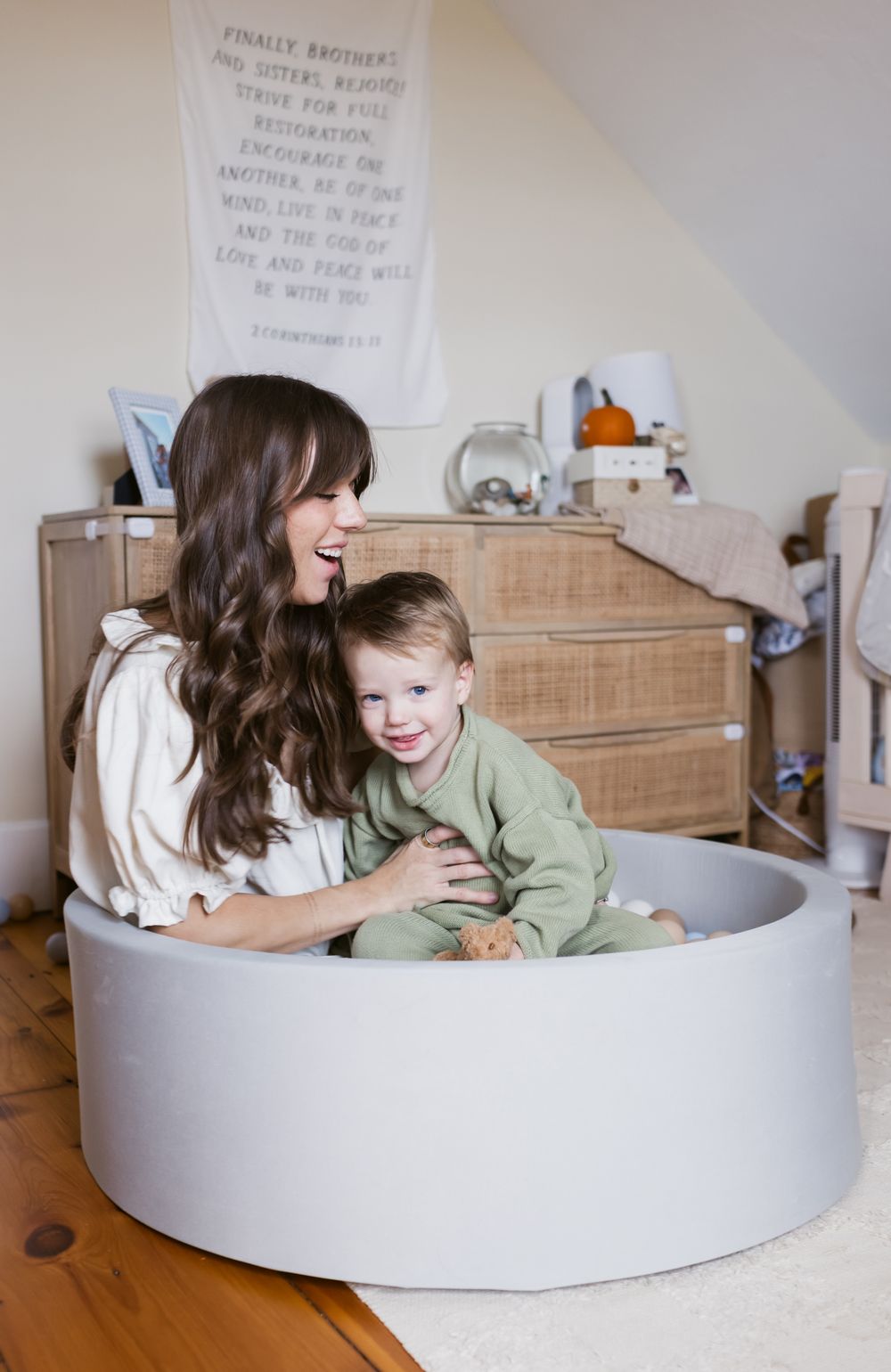 A mother with long brown hair sits with her toddler son in a foam ball pit in bedroom with a cloth sign on the wall for family photos