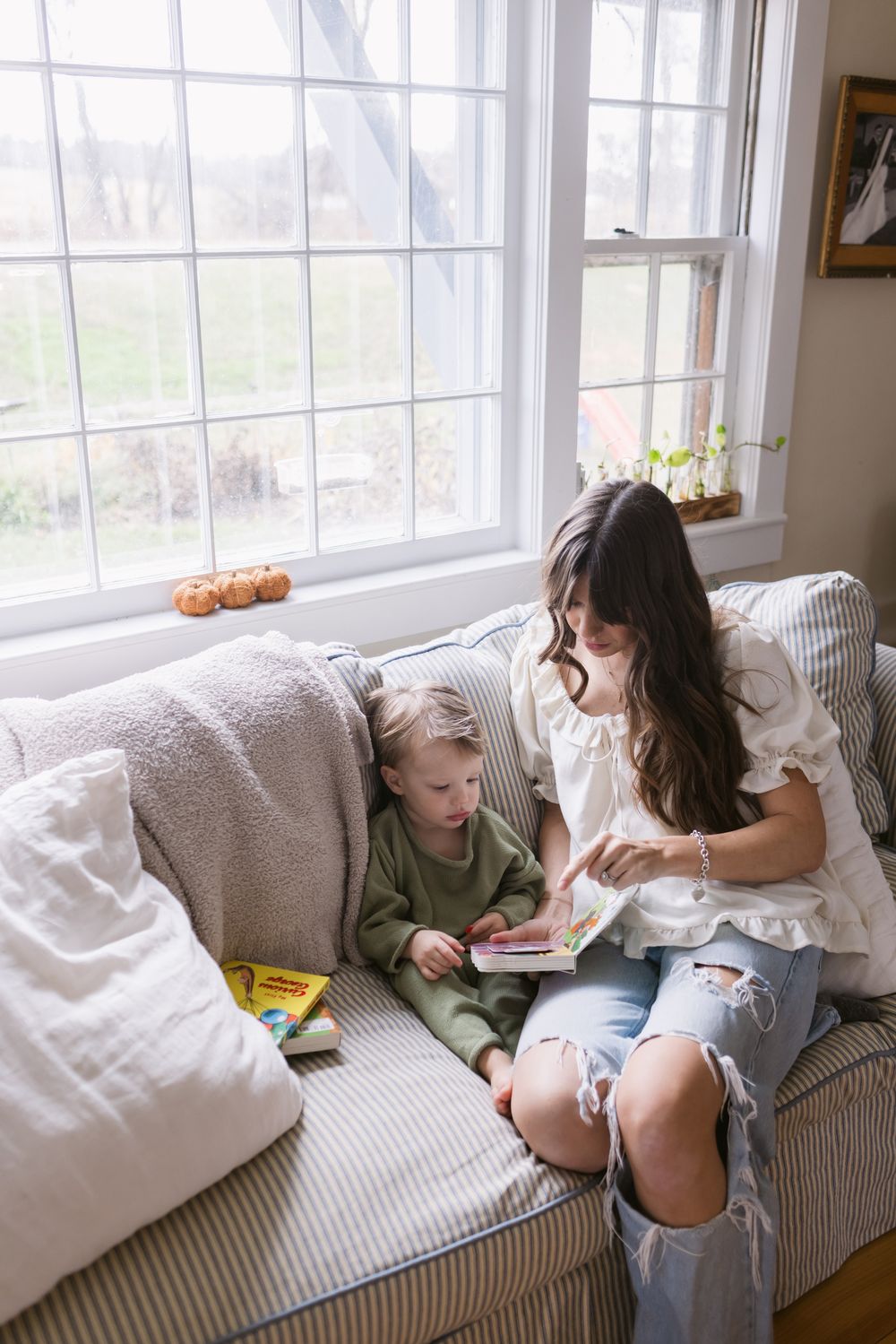 A mother with long brown hair and ripped jeans sits next to her toddler son reading books on a couch next to a large window in Harvard, MA