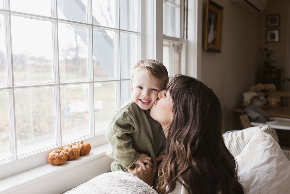 A brown haired mother kisses her toddler son on cheek sitting on a couch next to a window in Harvard, MA for lifestyle family photos
