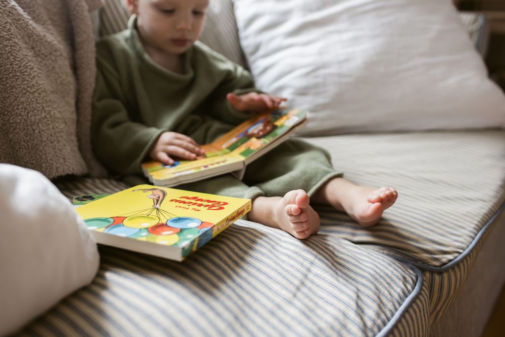 A toddler boy wearing sage sweater outfit sits on a couch reading books with a close up view of his toes for family in-home lifestyle photos