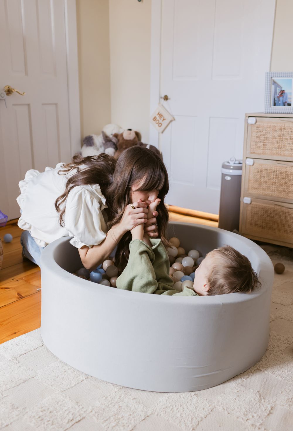 A mother with long brown hair playfully kisses her toddler son's feet while sitting in a foam ball pit in bedroom for family photos.