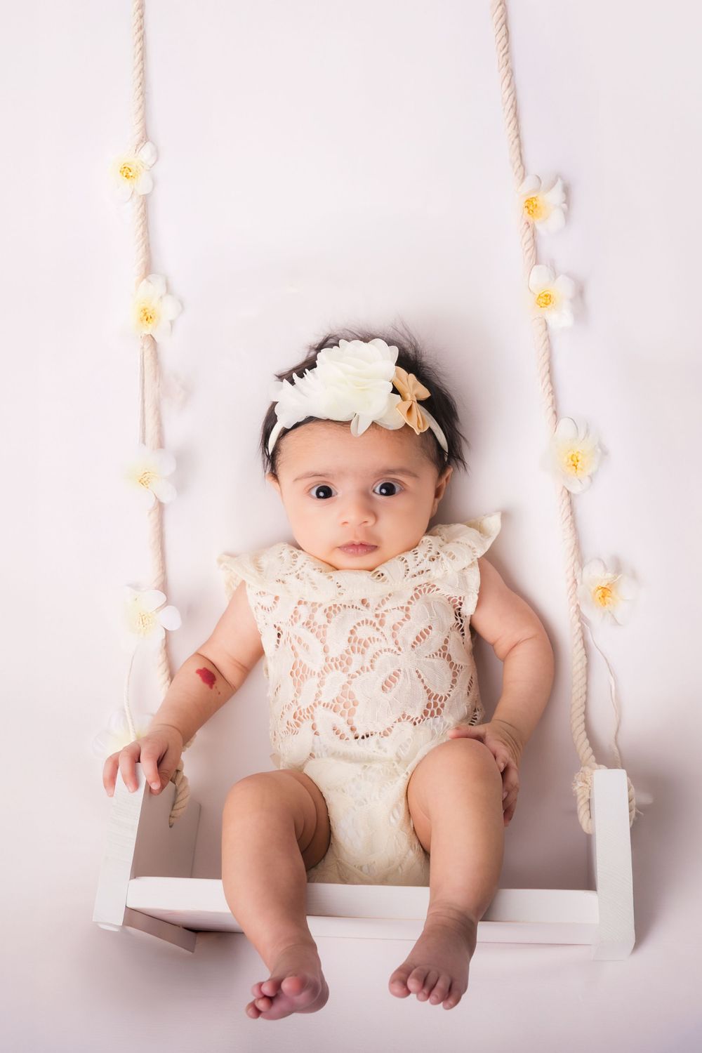 Baby in white lace romper sitting on decorative white swing with floral accents.
