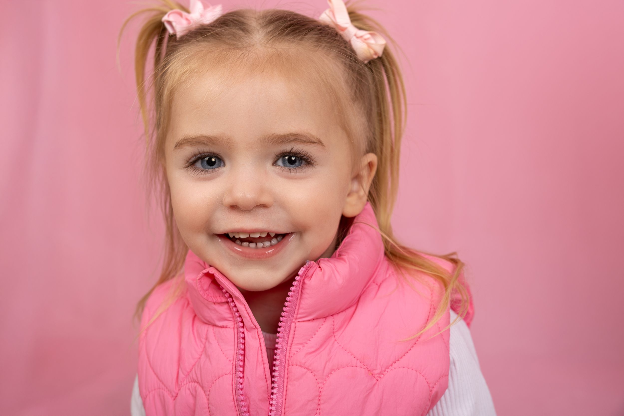 Child wearing pink puffy vest smiles against pink background.