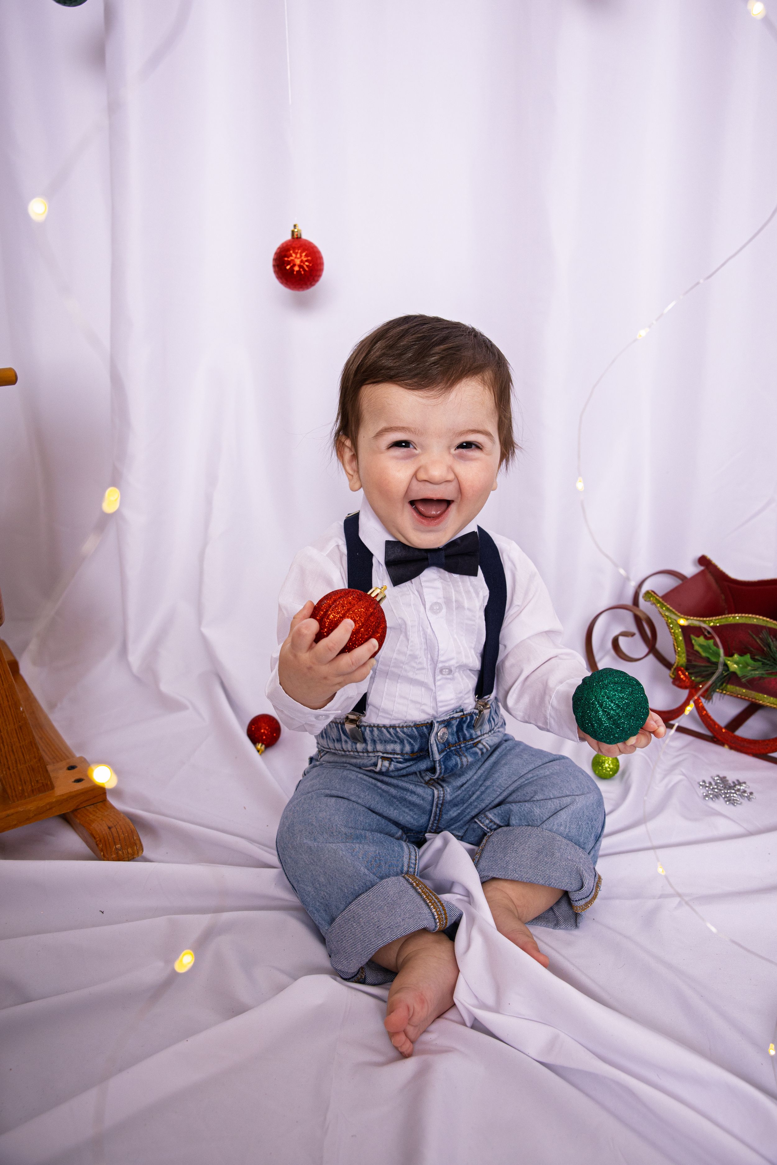 Baby in white shirt and jeans sits on white backdrop with festive Christmas ornaments.