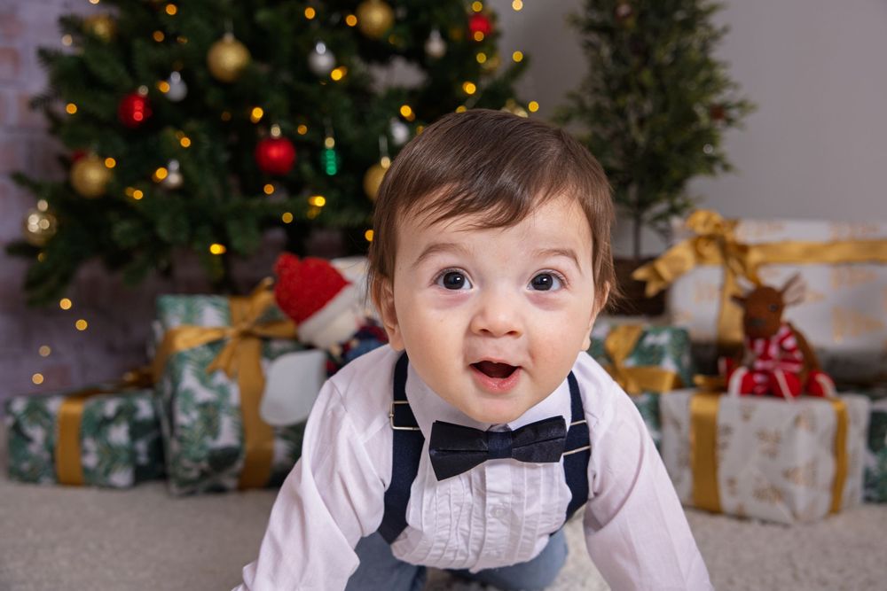 Baby in formal wear smiles near decorated Christmas tree and wrapped presents.