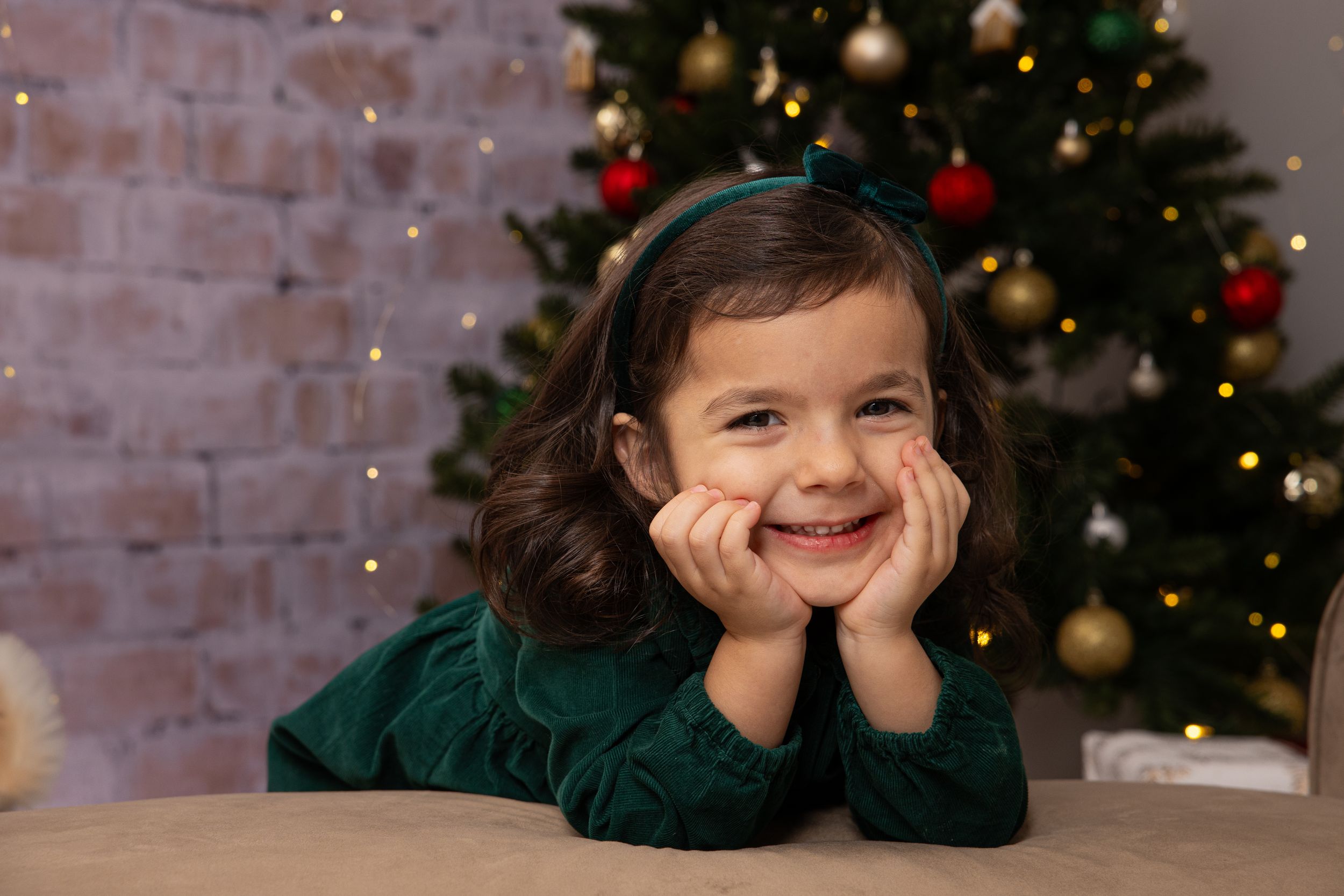 Child in green sweater poses playfully near Christmas tree with holiday decorations.