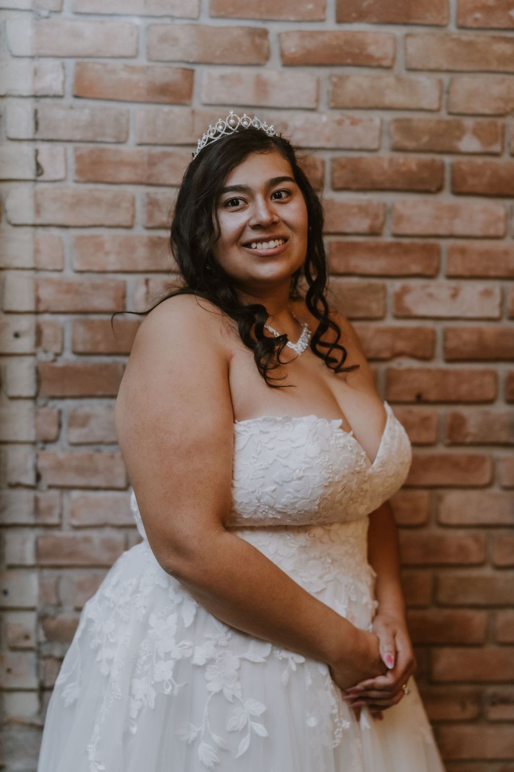 Bride in white lace dress poses against rustic brick wall.