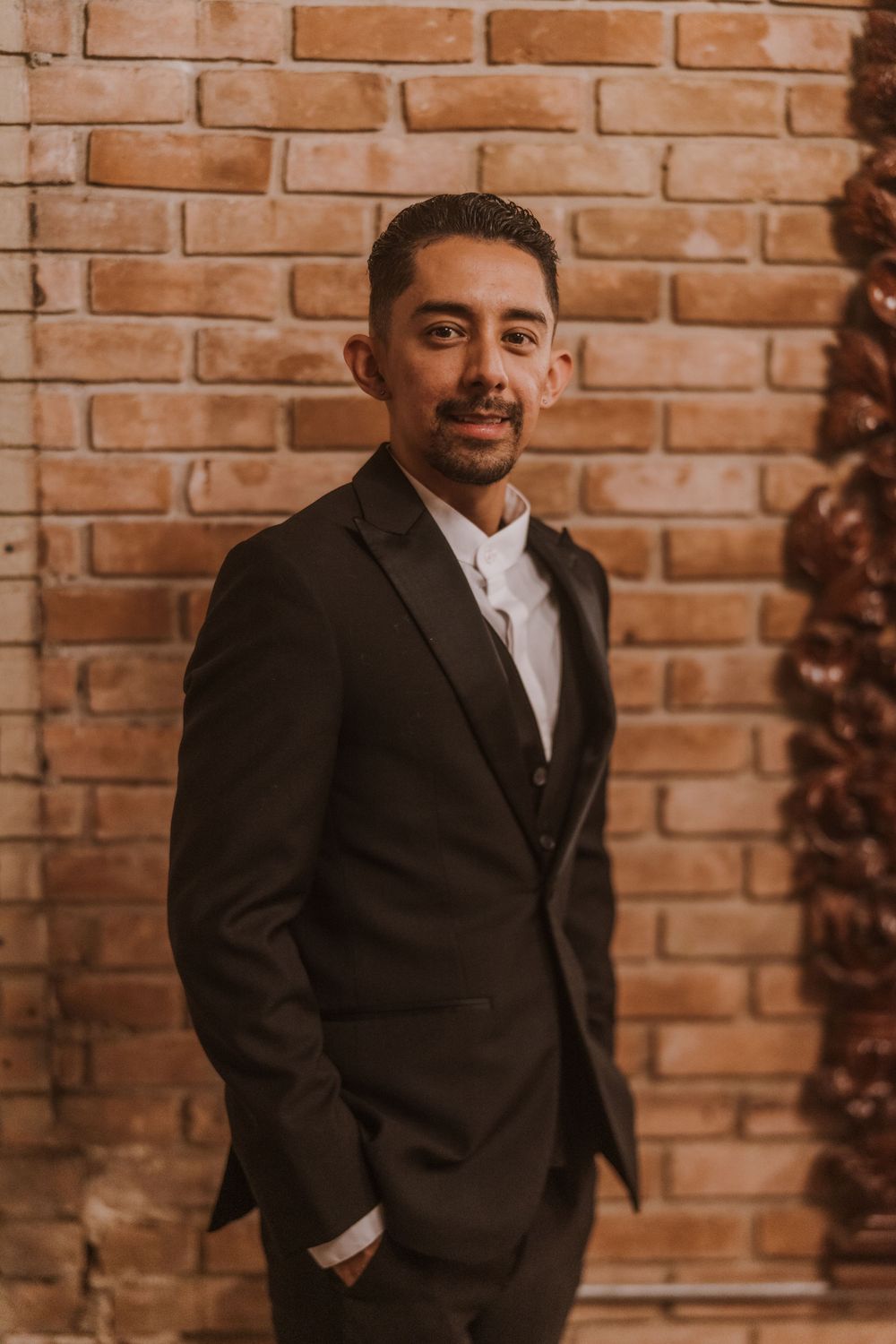 Groom in classic black suit poses against brick wall.