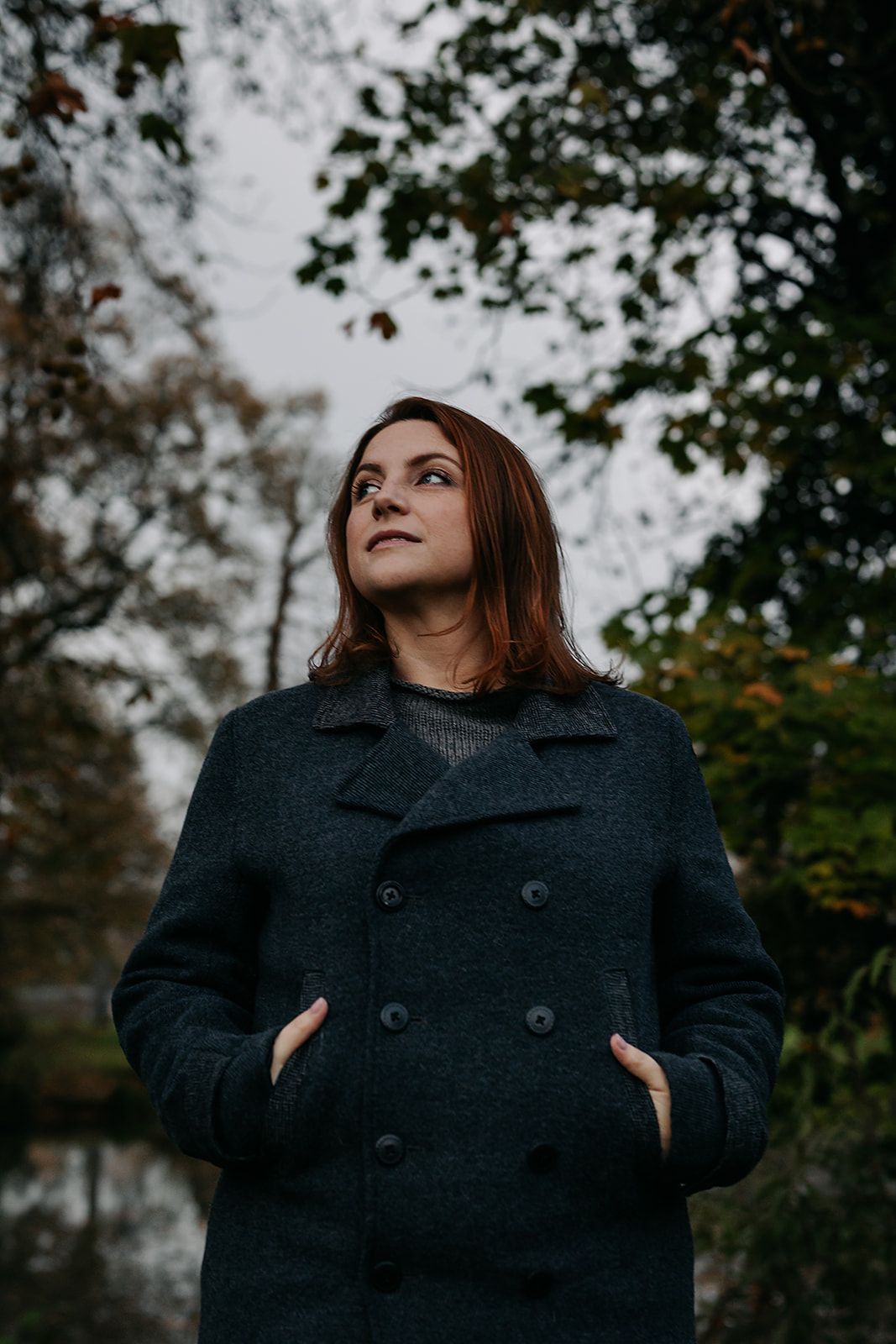 Woman in a dark wool peacoat poses thoughtfully against an autumn forest background.
