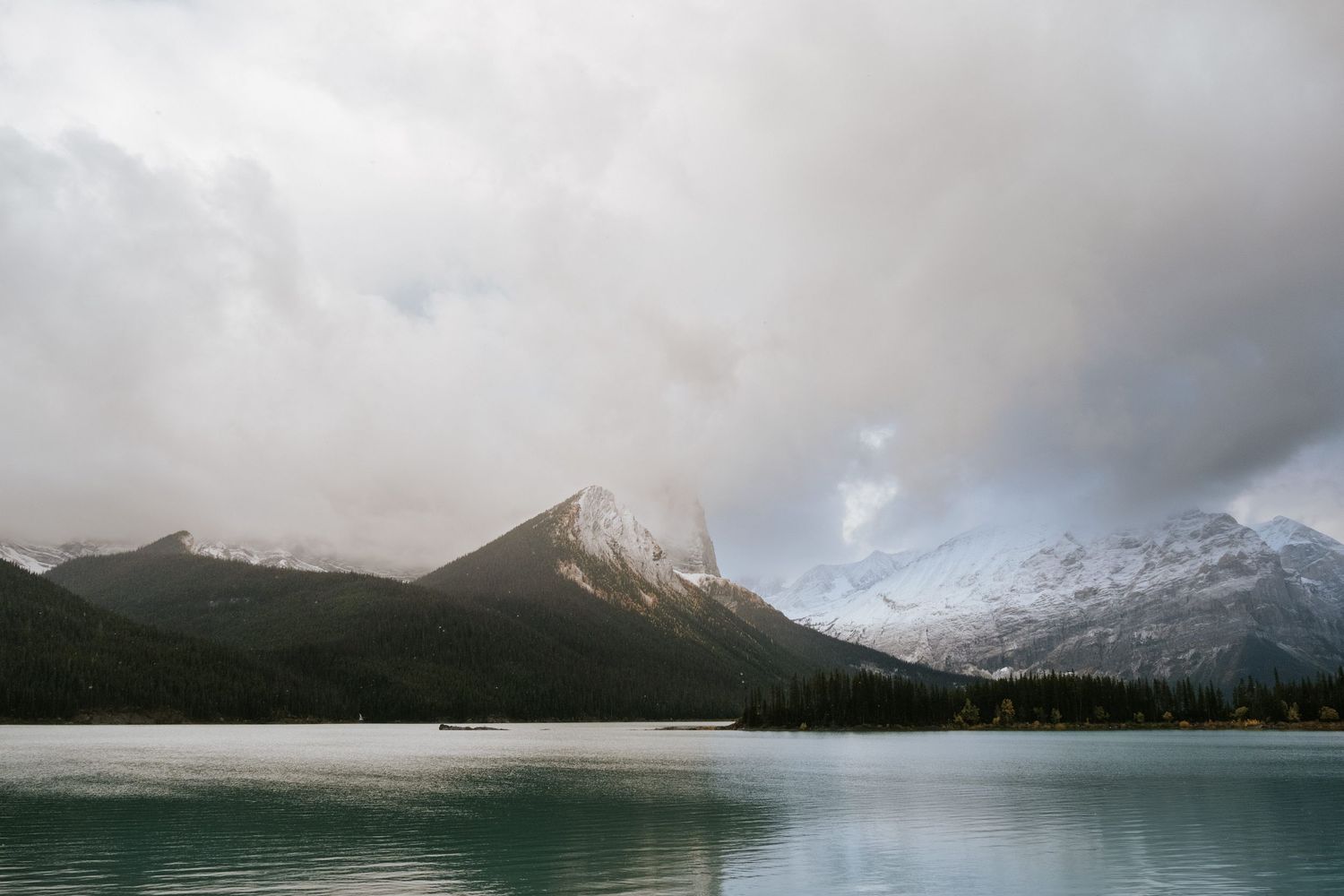 A scenic mountain lake reflects snow-capped peaks and dramatic clouds in the Canadian Rockies during sunset.