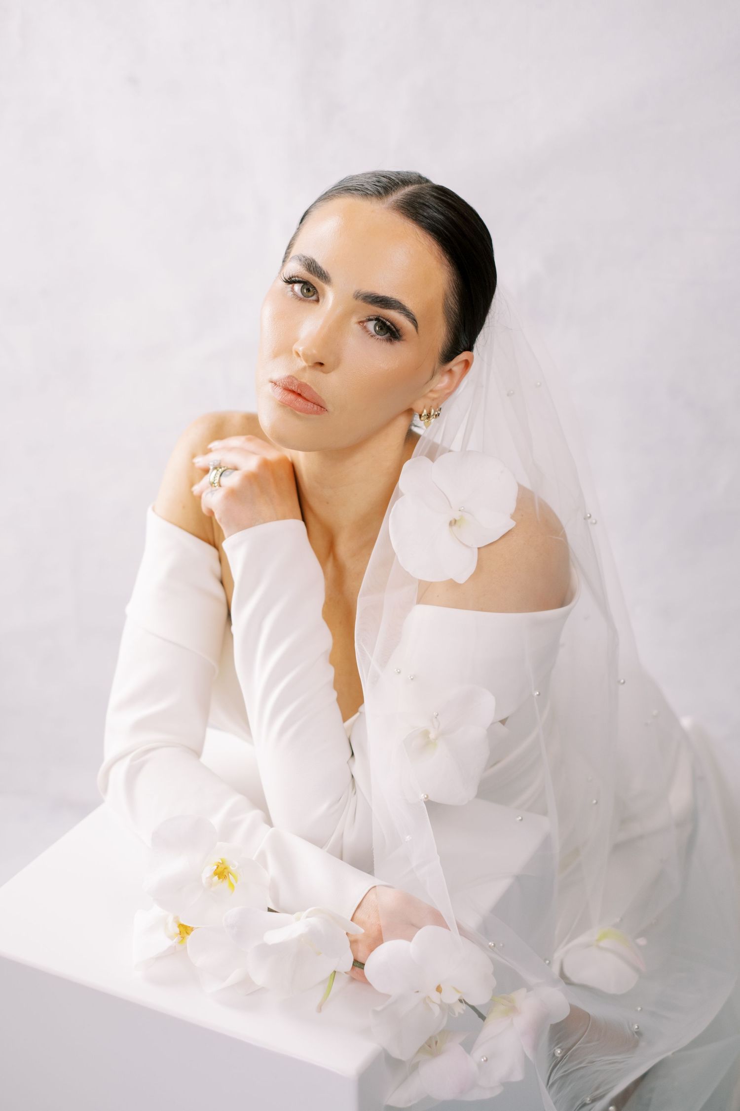 Elegant bridal portrait with white orchids and delicate veil against light background in soft natural lighting.