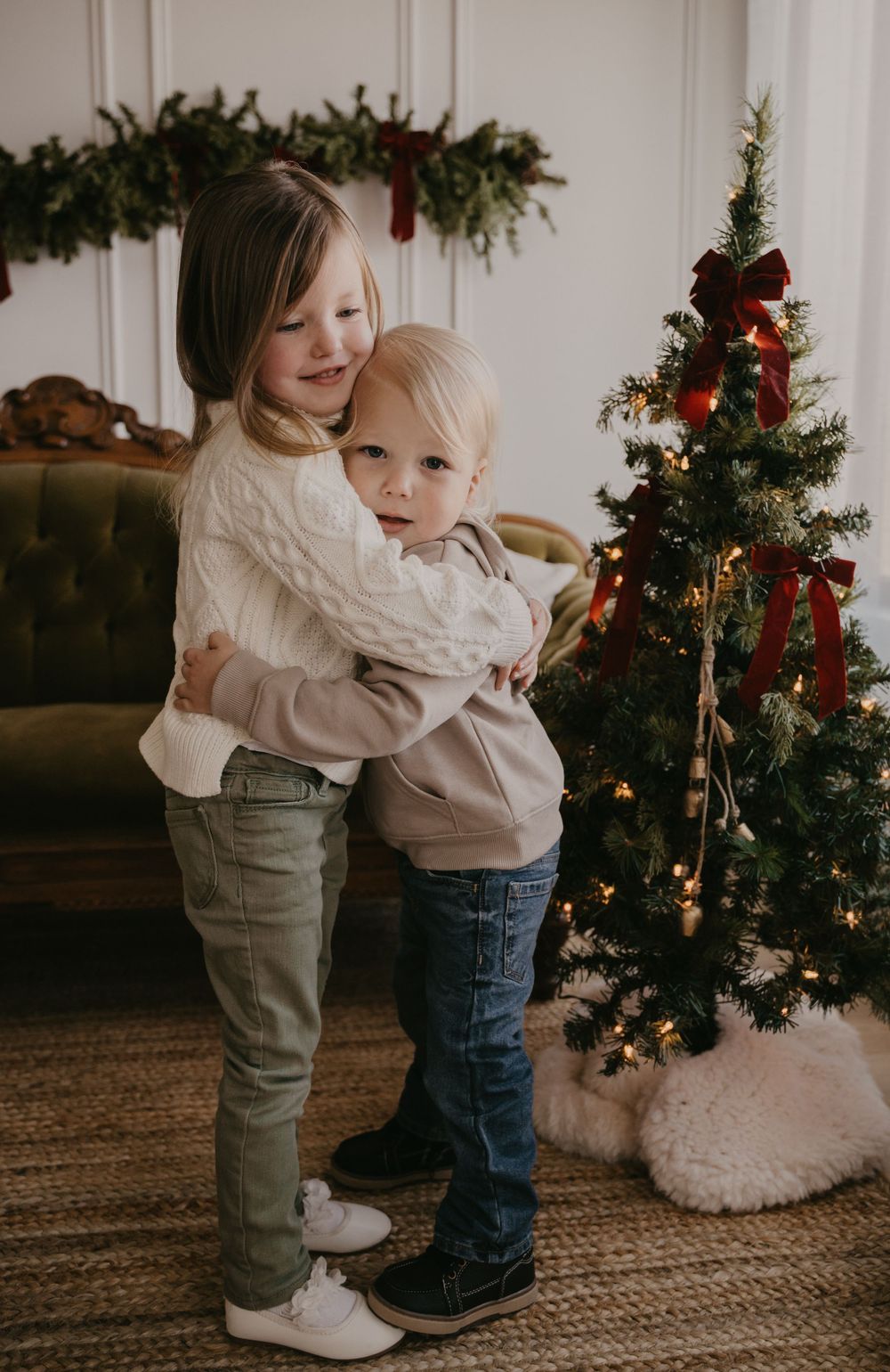 Two small children in matching outfits hug and play in front of a decorated Christmas tree on a textured rug.