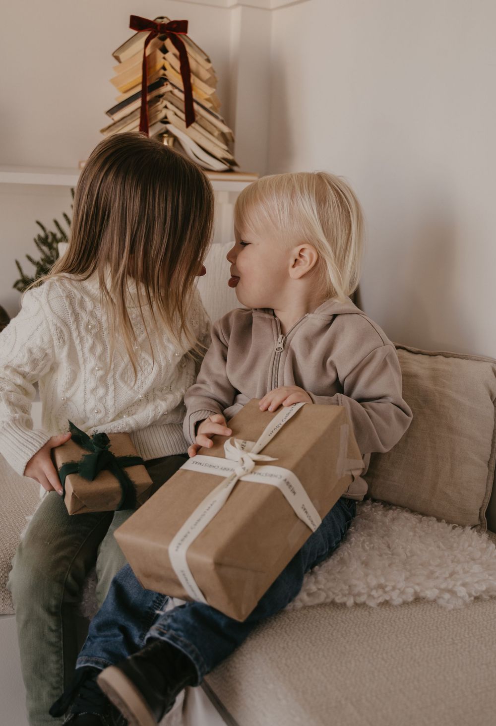 Two children sit together on a white carpet while exchanging wrapped presents in a minimalist room.