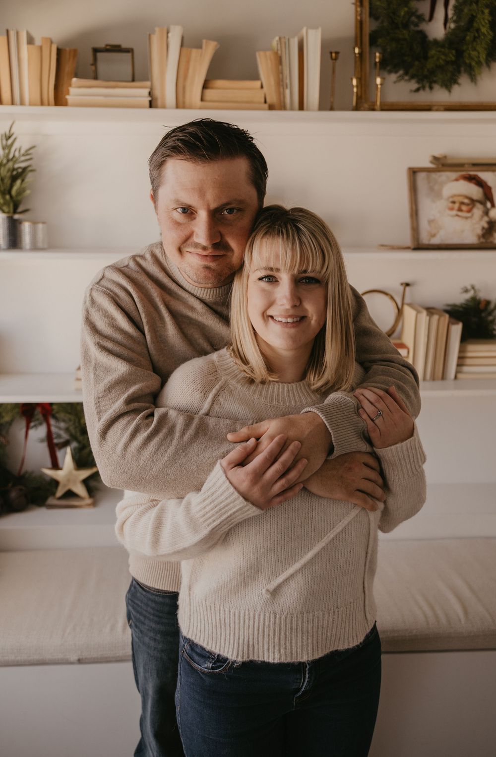 A couple in matching beige sweaters embrace in front of white shelves decorated with books and holiday decor.