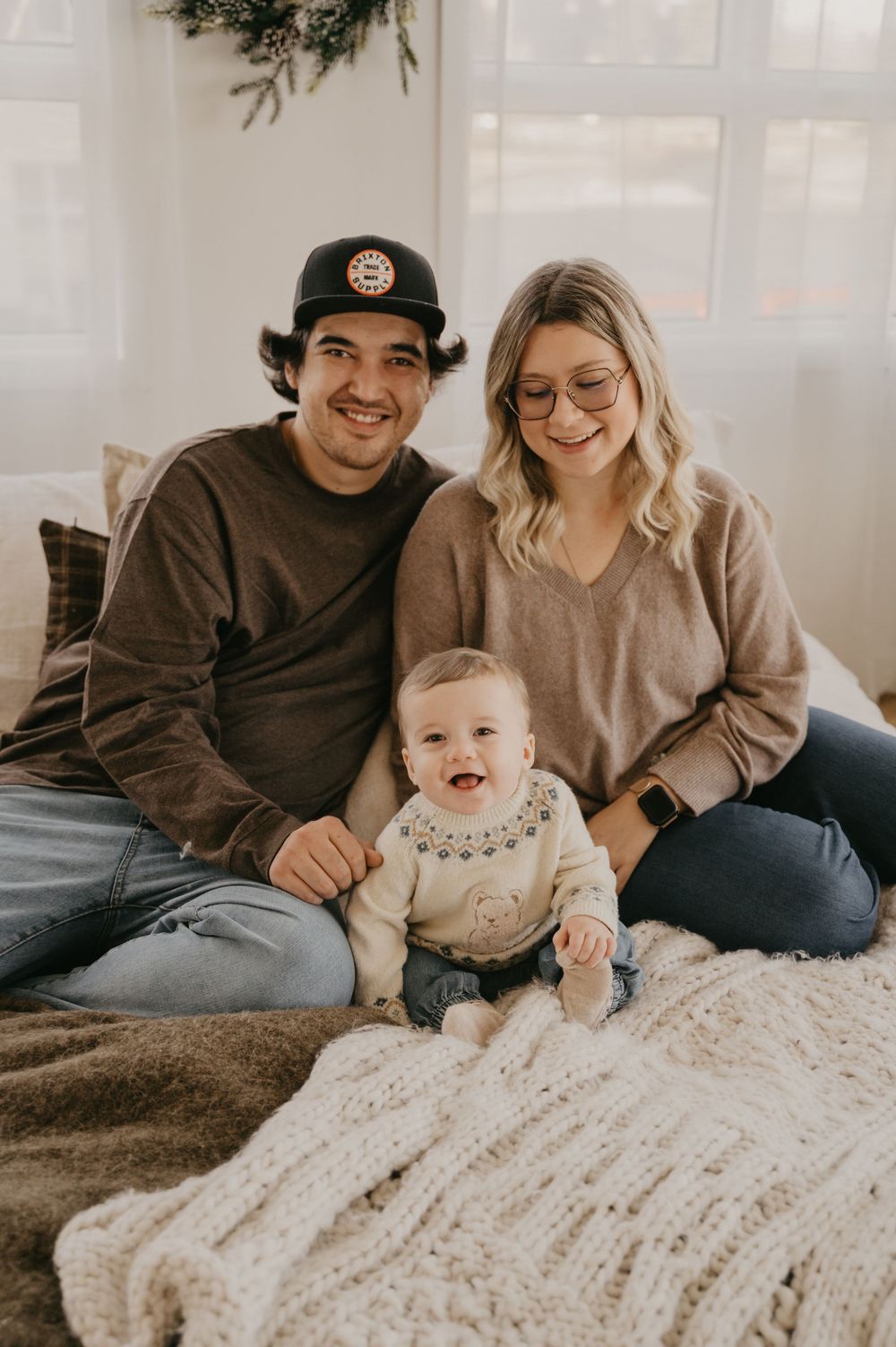 A family sits together on a cozy bed with white textured bedding in a bright room with a Christmas tree.