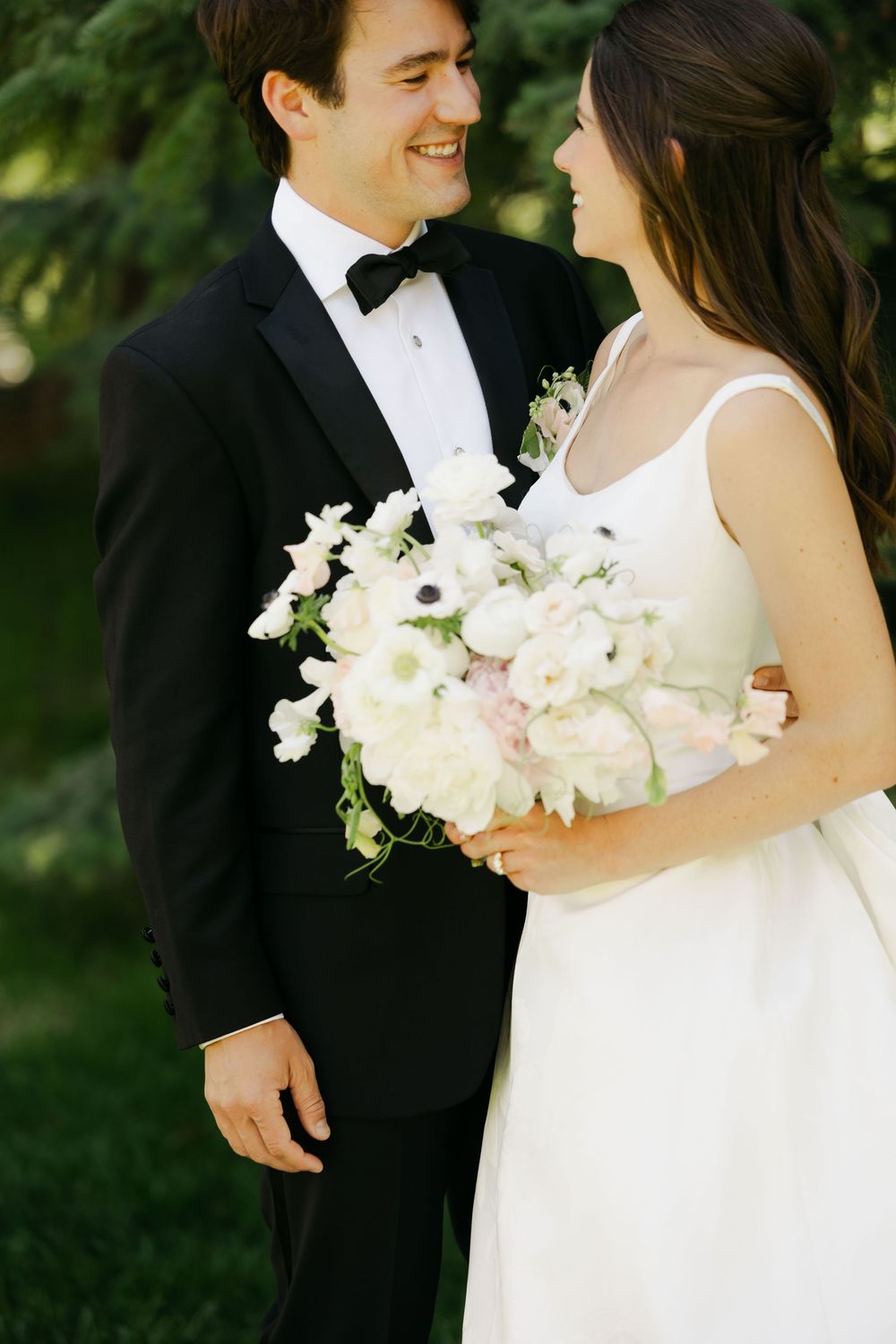 Elegant wedding couple in black tuxedo and white gown pose with white floral bouquet in outdoor garden setting.