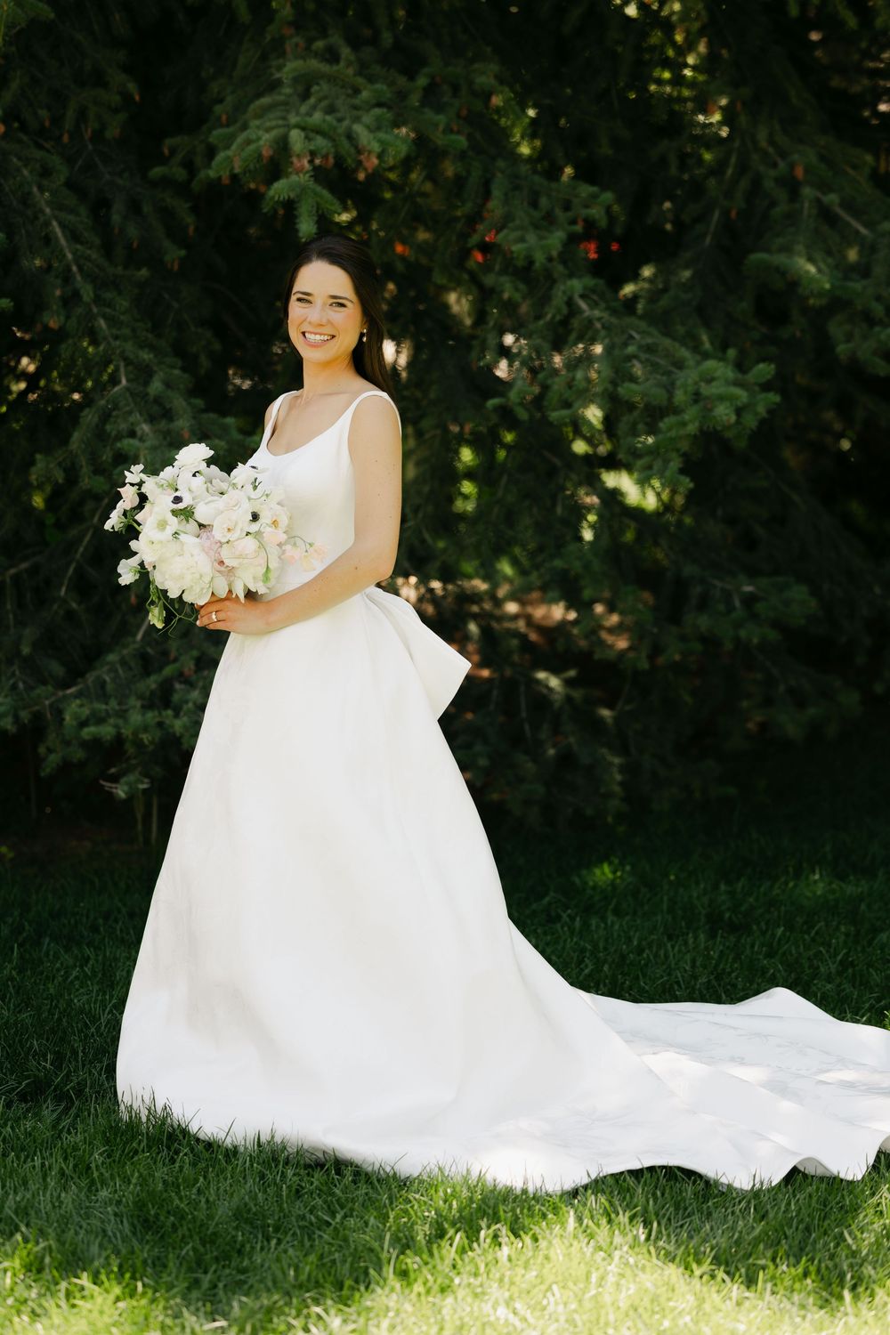 A bride in an elegant white ballgown poses gracefully on green grass with a white floral bouquet.