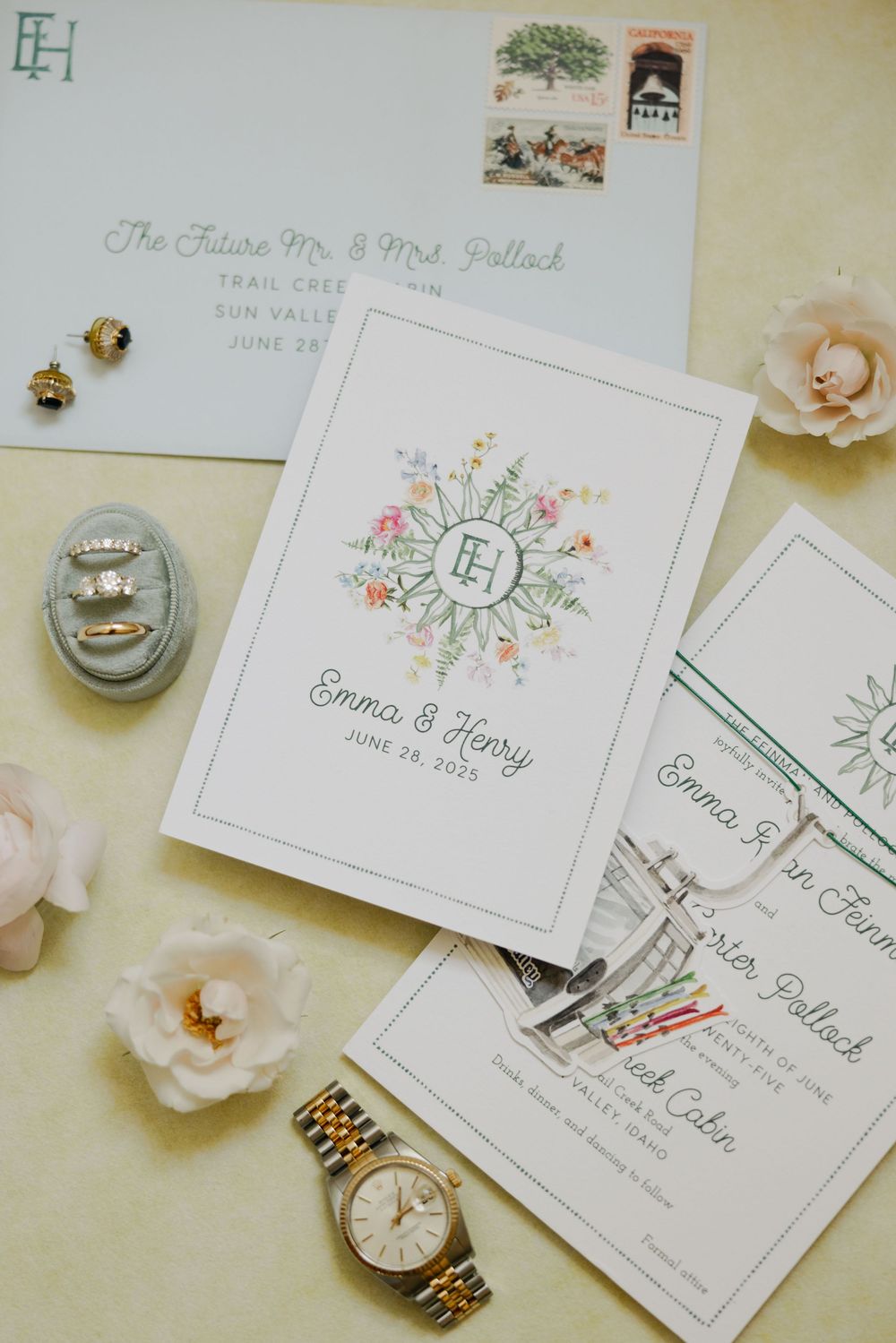Wedding invitation cards with floral designs displayed alongside jewelry and decor items on a cream colored surface.