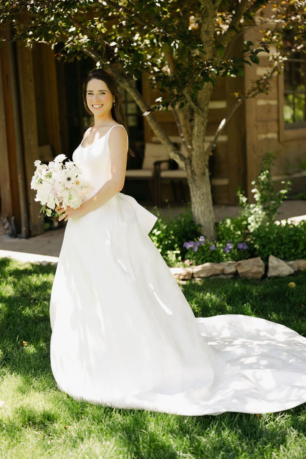 Flowing white wedding dress with long train photographed in a sunlit garden setting.