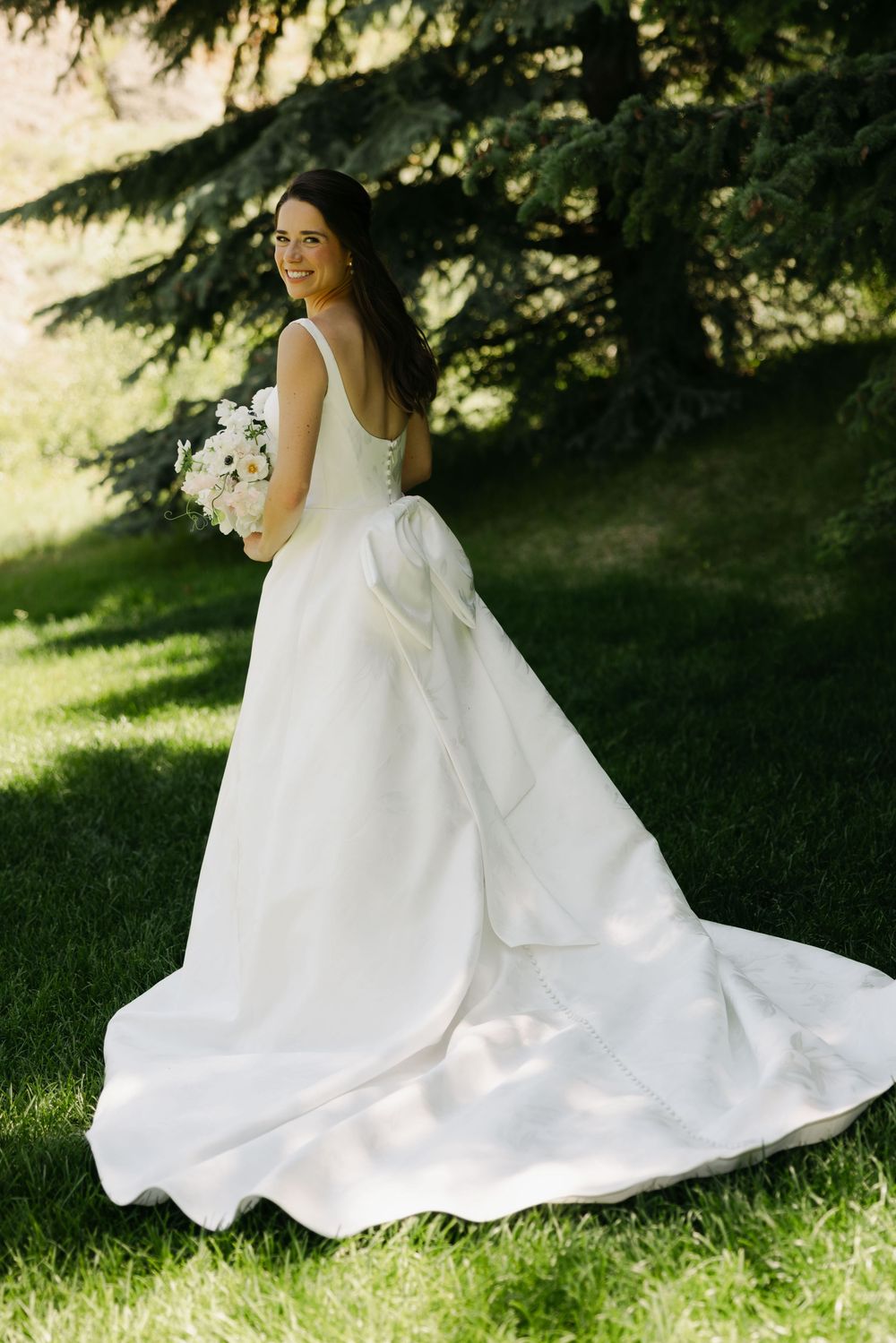 Bride in flowing white gown sitting gracefully on grass under evergreen trees.