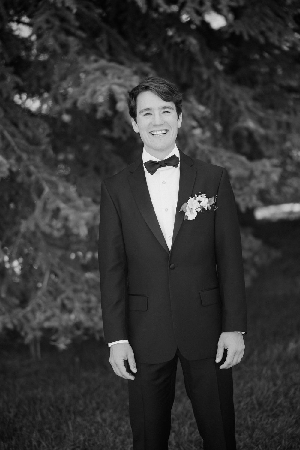 A groom in a classic black tuxedo and white bow tie smiles during an outdoor wedding photo session.