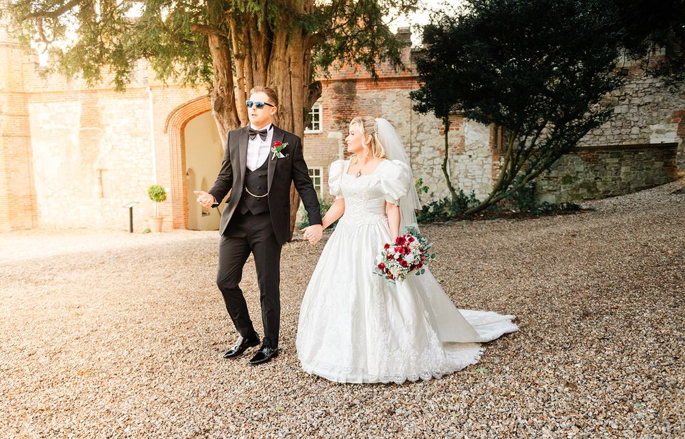 A bride and groom walk hand in hand on a gravel path surrounded by trees during golden hour.