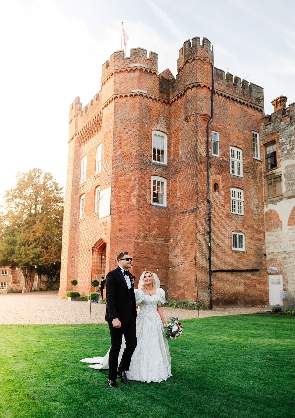 A couple in formal attire poses together on a green lawn in front of a historic red brick castle at sunset.