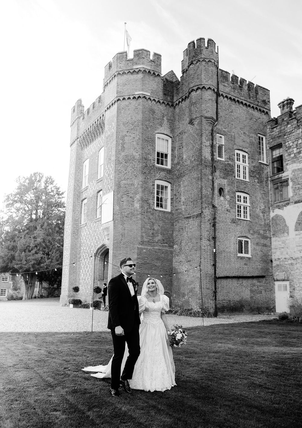 A wedding couple poses for romantic photos outside a majestic stone castle with turrets and towers in black and white.