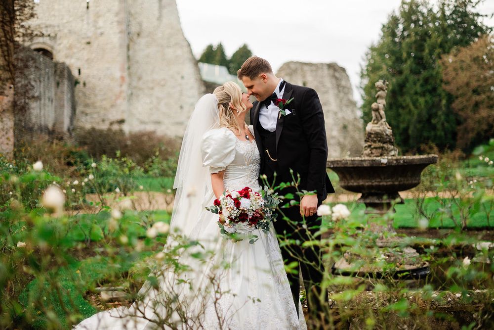 A romantic wedding couple embraces against a rustic stone wall backdrop with lush greenery.