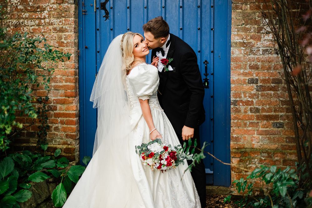 A romantic wedding couple shares a tender moment against a bright blue door and brick wall while holding a colorful bouquet.