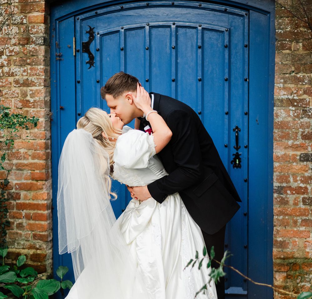 A couple shares a tender moment in front of a vibrant blue door with exposed brick walls.