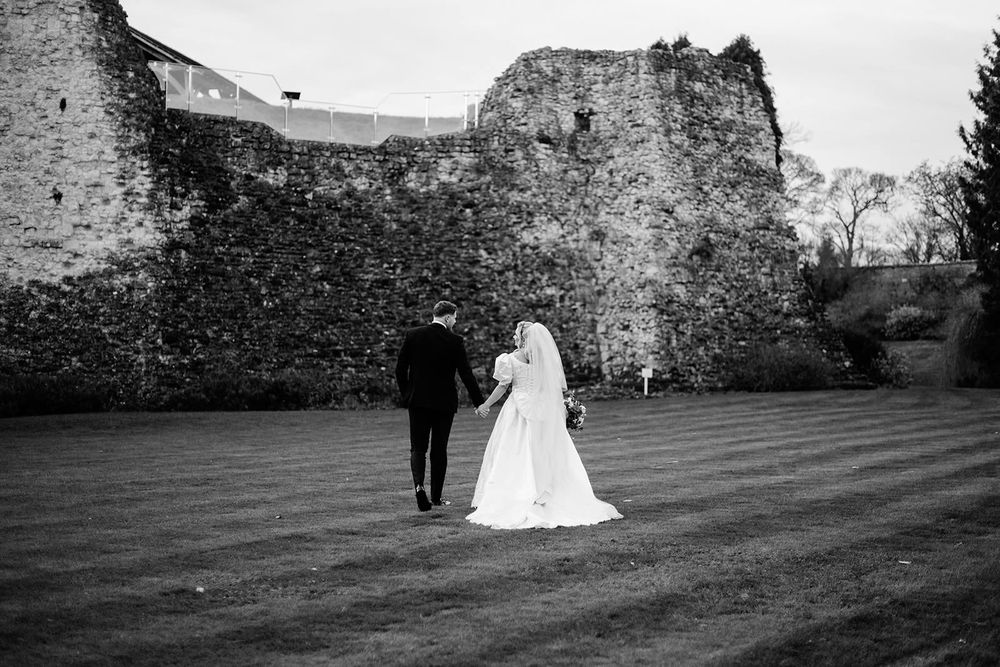 A couple walks across a grassy hillside near a historic stone castle ruin during a wedding photo session.