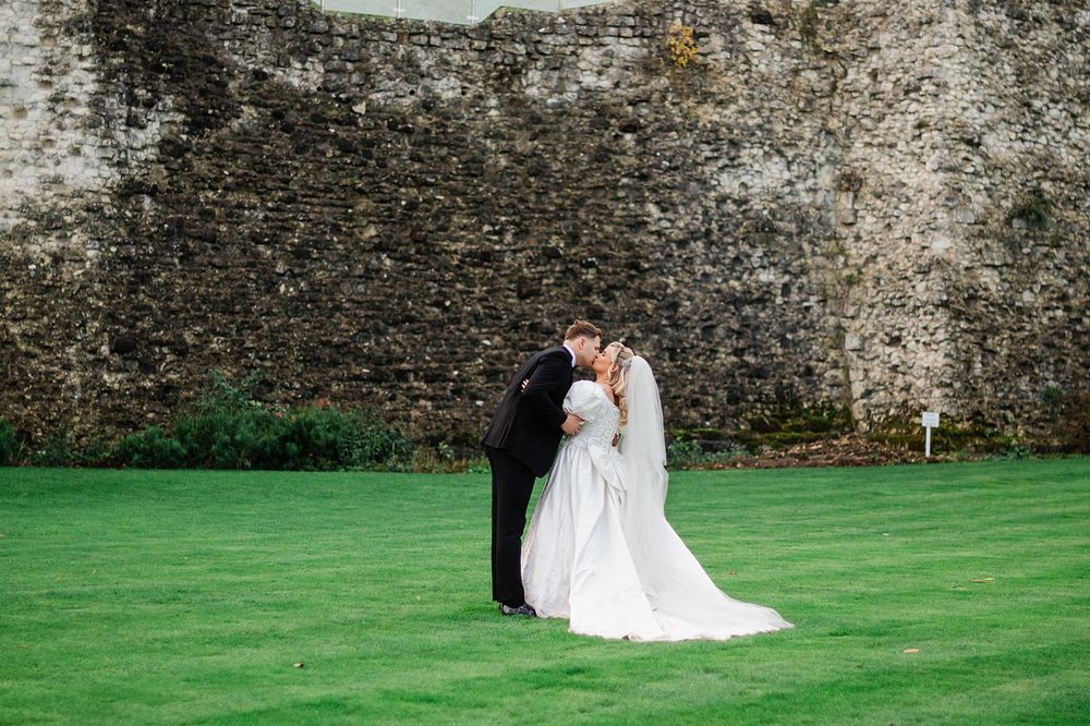Romantic couple in wedding attire sits together on lush green lawn against rustic stone wall backdrop.