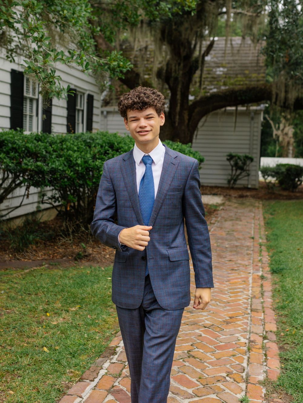 Person in a tailored navy plaid suit poses on brick pathway in garden setting.