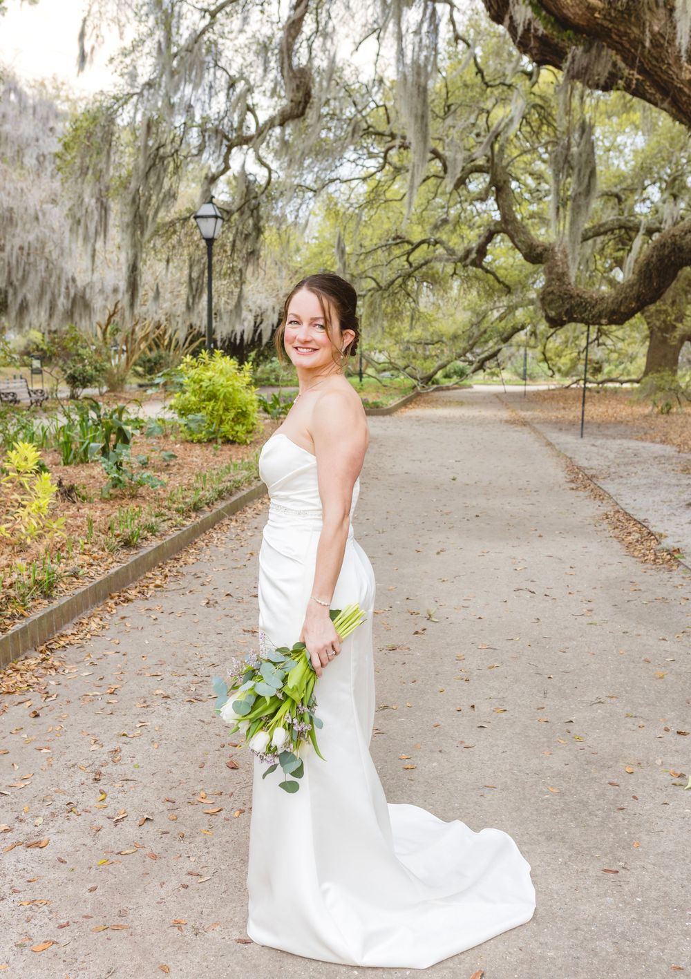 A bride in a white wedding dress poses on a tree-lined path with Spanish moss and spring foliage.